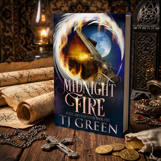 A book titled "Midnight Fire" by TJ Green stands upright on a rustic wooden table, surrounded by themed props including coins, a cross, scrolls, jewelry, and lanterns. The cover art shows a glowing fire, a coffin, a sword, and a full moon.